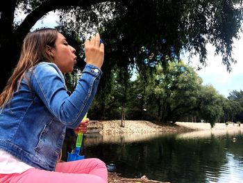 Woman blowing bubbles while sitting by lake against trees