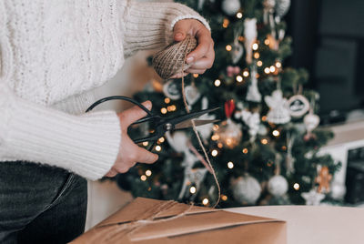 Midsection of woman wrapping christmas present in kraft paper in front of christmas tree at home