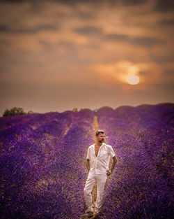 Full length of mid adult man standing on field amidst flowers against cloudy sky during sunset