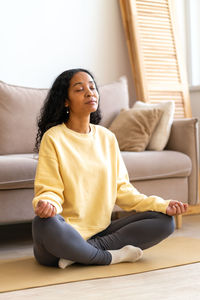 Young african-american female sitting in lotus yoga pose on mat in while meditating, vertical