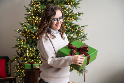 A smiling girl with glasses unties a burgundy bow on a green box near the christmas tree