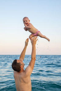 Shirtless man throwing baby girl mid-air while standing against sky in sea