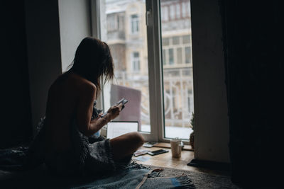 Woman using mobile phone while sitting on window