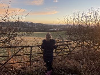 Woman standing by bare tree against sky during sunset