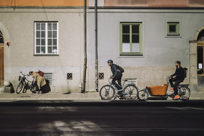 Male commuters riding bikes on road against building during sunny day