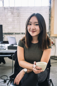 Portrait of smiling female computer programmer holding coffee cup while sitting at creative office