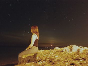 Side view of woman sitting on rock against sky at night