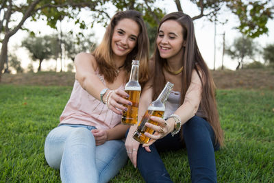 Smiling friends with alcoholic drinks in bottle sitting on grassy field at park