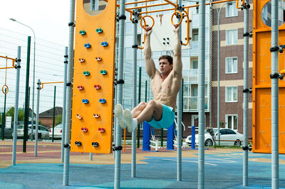 Shirtless young man exercising on gymnastic rings against building