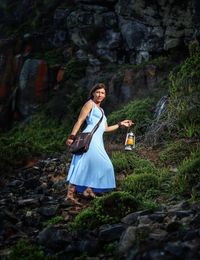 Full length of woman holding lantern while walking on rock in forest