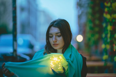 Thoughtful beautiful woman with blue scarf and glowing jar in city at dusk