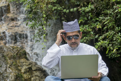 Mature man wearing sunglasses while using laptop against waterfall