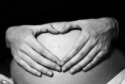 Midsection of pregnant woman making heart shape on abdomen against black background