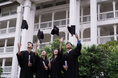 Students in university gowns throwing mortarboards while standing against building