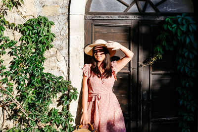 Beautiful young woman in pink dress standing in front of picturesque old house, door, light, shadow.