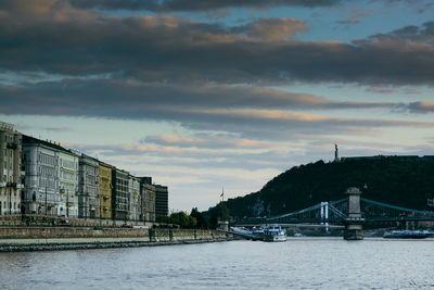 Bridge over river by buildings against sky at sunset