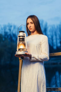 Young woman with illuminated oil lamp standing in forest at dusk