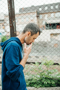 Side view of young man standing by chainlink fence