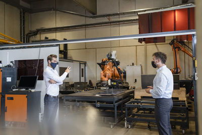 Businessmen with face mask discussing while standing at social distance against robotic arm equipment in factory