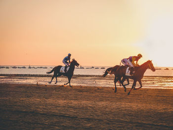 People riding horse on beach against sky during sunset