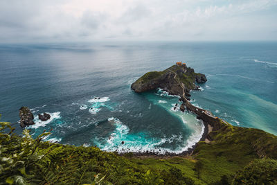 Drone view of paving stone way leading along stone bridge and ridge of rocky hill to lonely house on island gaztelugatxe surrounded by tranquil sea water under cloudy sky in basque country