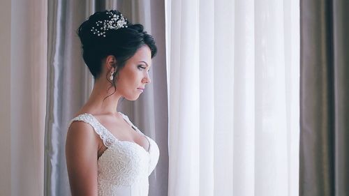 Thoughtful bride standing by curtain at home