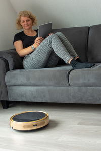 Woman holding digital tablet while sitting on sofa at home by vacuum cleaner