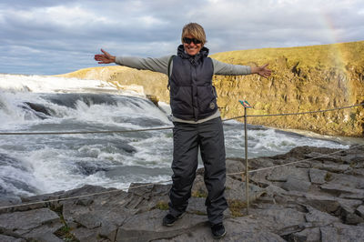 Smiling woman with arms outstretched standing on rock by sea against sky