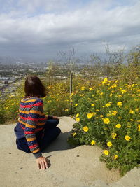 Side view of woman sitting by yellow flowering plants against cloudy sky