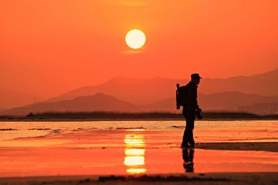 Silhouette man standing on shore at beach against orange sky