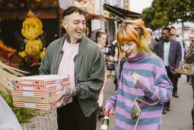 Happy non-binary person holding pizza boxes while walking with friend on sidewalk in city