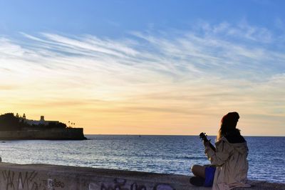 Rear view of woman photographing sea against sky during sunset