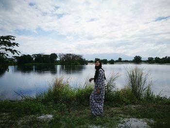 Woman looking up while standing on field by lake against cloudy sky