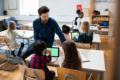 Teacher sitting on desk while explaining students using digital tablet in classroom