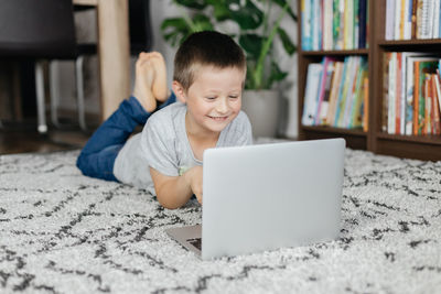 Charming schoolboy boy lies on the floor and watches lessons or chats online with friends. 