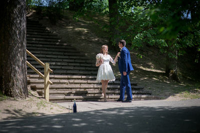 Full length of wedding couple holding hands on steps at park during sunny day