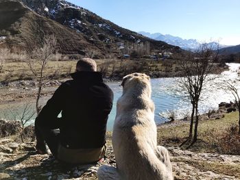 Rear view of man sitting with dog at riverbank against mountain