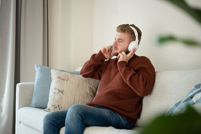 Young caucasian blond man listening to music with big white handsfree headphones sitting on sofa