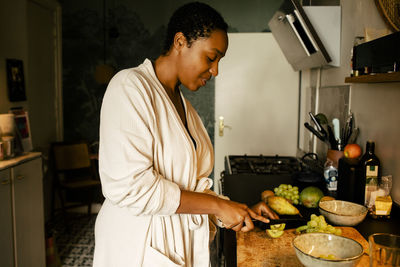 Side view of young woman cutting kiwi at kitchen counter