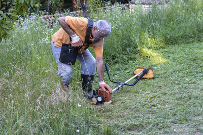 A mature man in protective clothing, and gloves  and mowing tall grass and the weeds with a trimmer