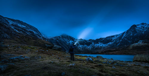 Man with flashlight standing at lakeshore against mountains