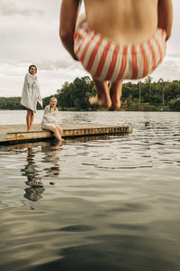 Women at jetty looking at male friend jumping in lake during vacation