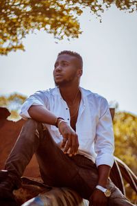 Young man looking away while sitting by tree against sky