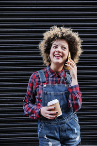 Portrait of smiling young woman using mobile phone while standing against wall