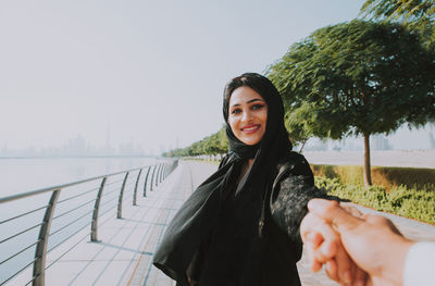 Portrait of smiling young woman standing by railing against sky