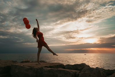 Woman holding balloons while standing on rock by sea against sky during sunset