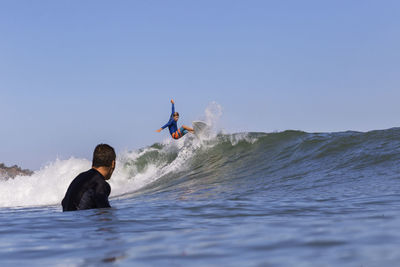 Man looking at friend surfing on sea against clear blue sky