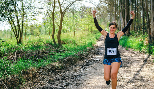 Cheerful woman with arms raised running in forest during marathon