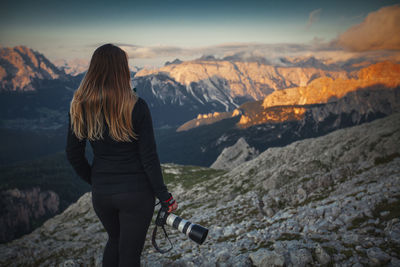Rear view of woman photographing while standing against mountains during sunset