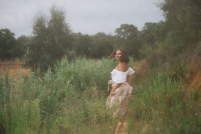 Side view of young woman standing amidst plants against sky in forest